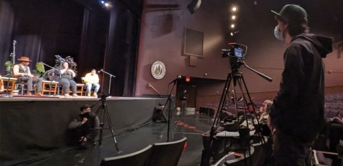 A student operates a video camera in the foreground with people seated on stage in the background