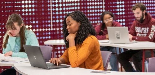 Students working at long tables on laptops with textbooks