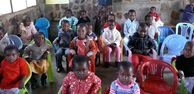 Little children sitting in chairs in classroom staring wide-eyed up at the camera