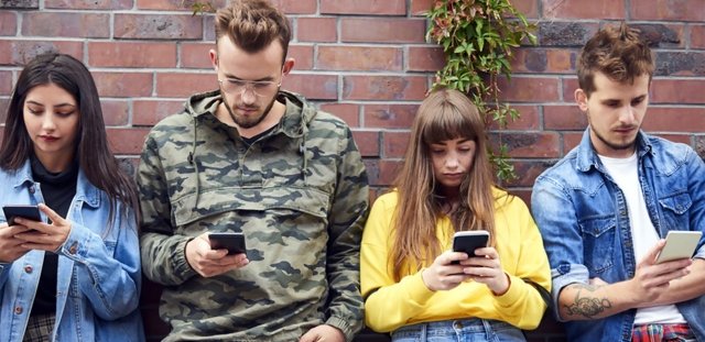 Four college students, leaning against the wall, staring at their phones